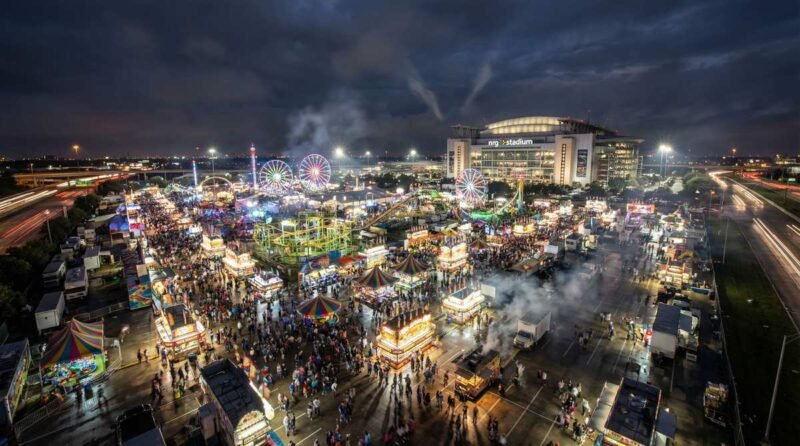 Aerial nighttime view of the Houston Rodeo carnival grounds at NRG Park, with colorful rides and a large crowd.